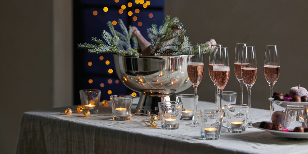 Elegancki, świąteczny st&oacute;ł udekorowany świeczkami w szklanych świecznikach, kieliszkami z r&oacute;żowym winem musującym oraz dużym metalowym coolerem wypełnionym lodem i gałązkami iglastymi. W tle widać rozmyte, ciepłe światełka dekoracyjne.